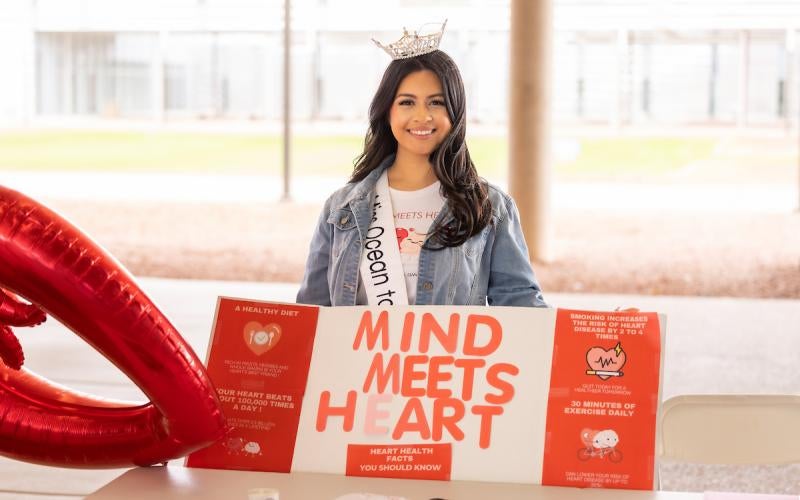 Miss Ocean to Ocean wears a sash and crown while holding a "Mind Meets Heart" poster with information about heart health at the 2025 Heart & Healthy Relationships Fair.