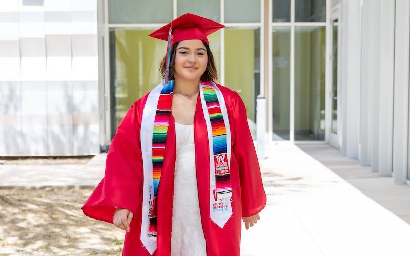 Student graduate walks toward camera in cap and gown outside of a building at AWC.