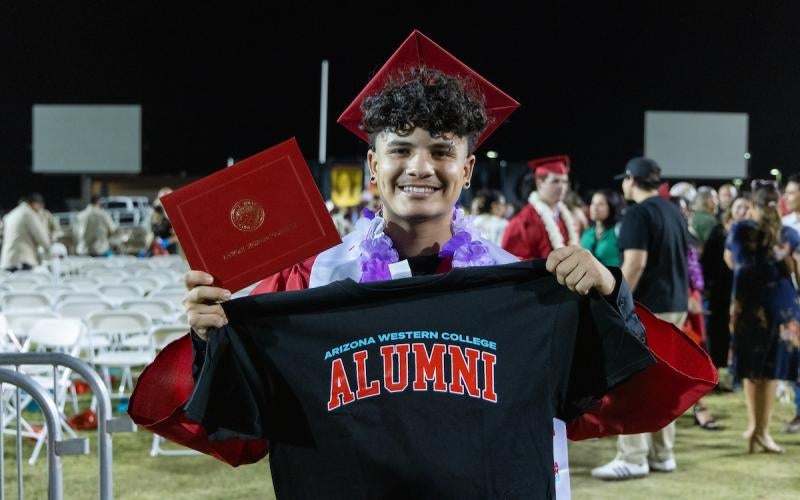 An AWC student in graduation regalia holds up an "Arizona Western College ALUMNI" T-shirt.