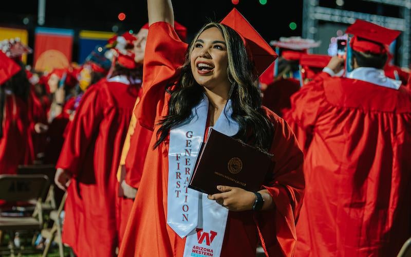 First Generation student in cap and gown smiles and holds up her diploma at the 2024 Commencement ceremony while fireworks go off behind her.