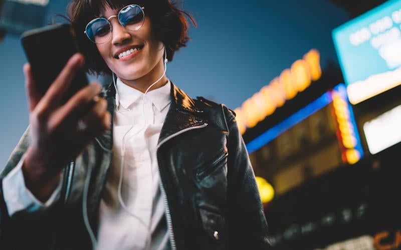 Stock image of a girl in a leather jacket with headphones in, smiling at her phone.